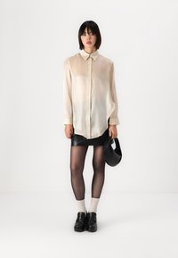Beige szatén ing gombos záródással és gallérral, fekete lakk mini szoknyával, áttetsző fekete harisnyával és fekete platform cipőkkel párosítva.