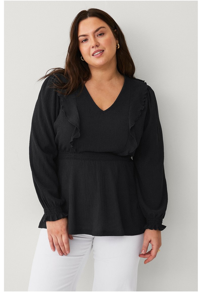 Blouse noire texturée avec des volants le long du décolleté en V et des épaules ; manches longues avec poignets élastiques, associée à un pantalon blanc.