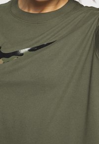 Camiseta verde oliva de algodón, con un logotipo negro brillante de Nike que presenta detalles de camuflaje. Cuello redondo, mangas cortas, textura suave.