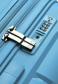 Valigia azzurra chiara con esterno testurizzato, doppie zip e lucchetto TSA con combinazione a tre cifre. Dettagli di cucitura visibili.