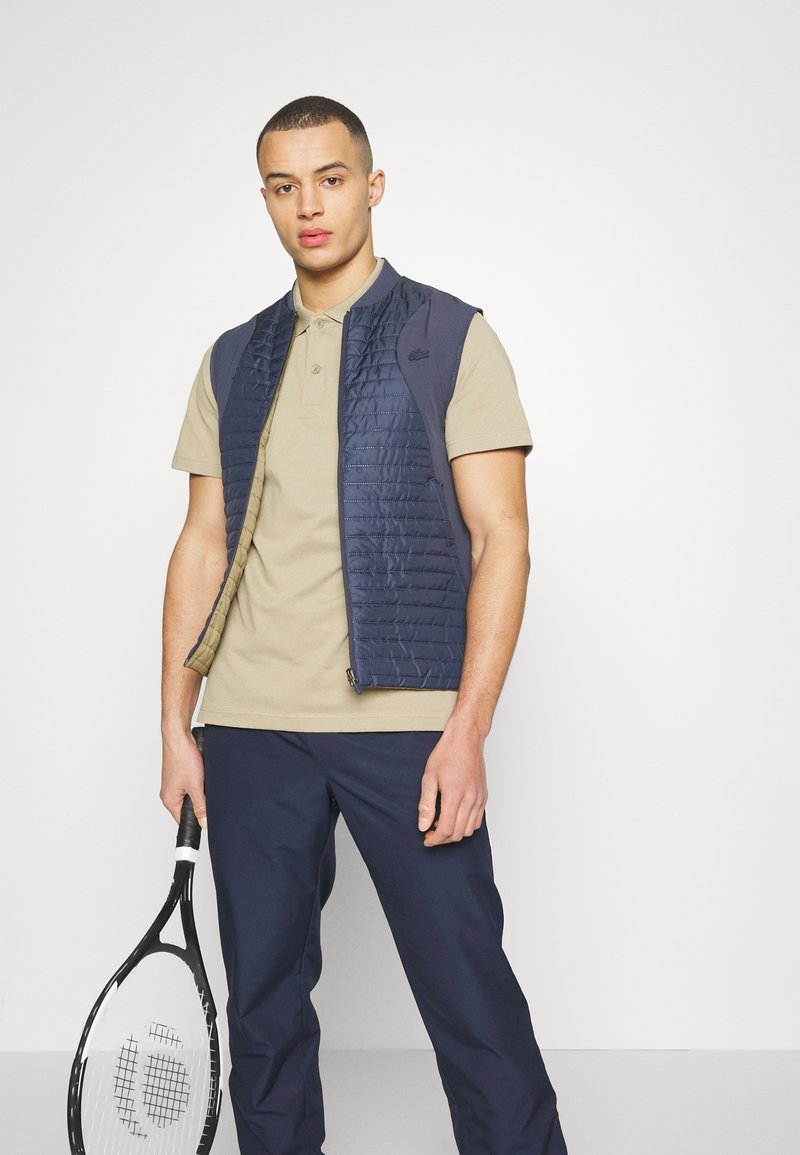 Gilet matelassé bleu marine sur une chemise polo beige, associé à un pantalon sombre, tenant une raquette de tennis. Le gilet dispose d'une fermeture éclair et de poches latérales.