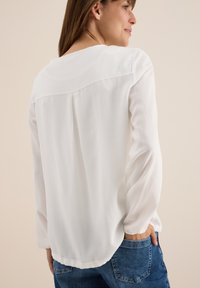 Blusa blanca con mangas largas ligeramente abullonadas y finas rayas verticales, con un escote redondeado y un corte holgado en la parte trasera.