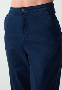 Marineblauwe broek van zacht materiaal, met een knoopsluiting aan de voorkant, zijzakken en een getailleerde pasvorm. Gladde textuur met minimale details.