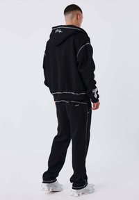 Hombre vestido con sudadera con capucha negra y pantalones a juego, con zapatillas blancas, de pie frente a un fondo claro y liso, mirando hacia atrás.