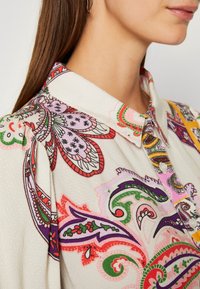 Chemise à col avec une base blanche ornée de motifs cachemire et floraux colorés. Le tissu semble texturé avec des boutons sur le devant.