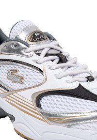 Sneaker bianche realizzate con materiali in rete e pelle, con accenti metallici argento e oro, sezioni testurizzate e un logo sulla linguetta.