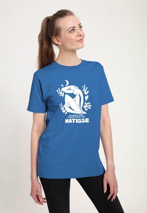 Ung kvinde med langt hår iført en blå Matisse kunstprint t-shirt og sorte bukser, der kigger let opad og smiler.