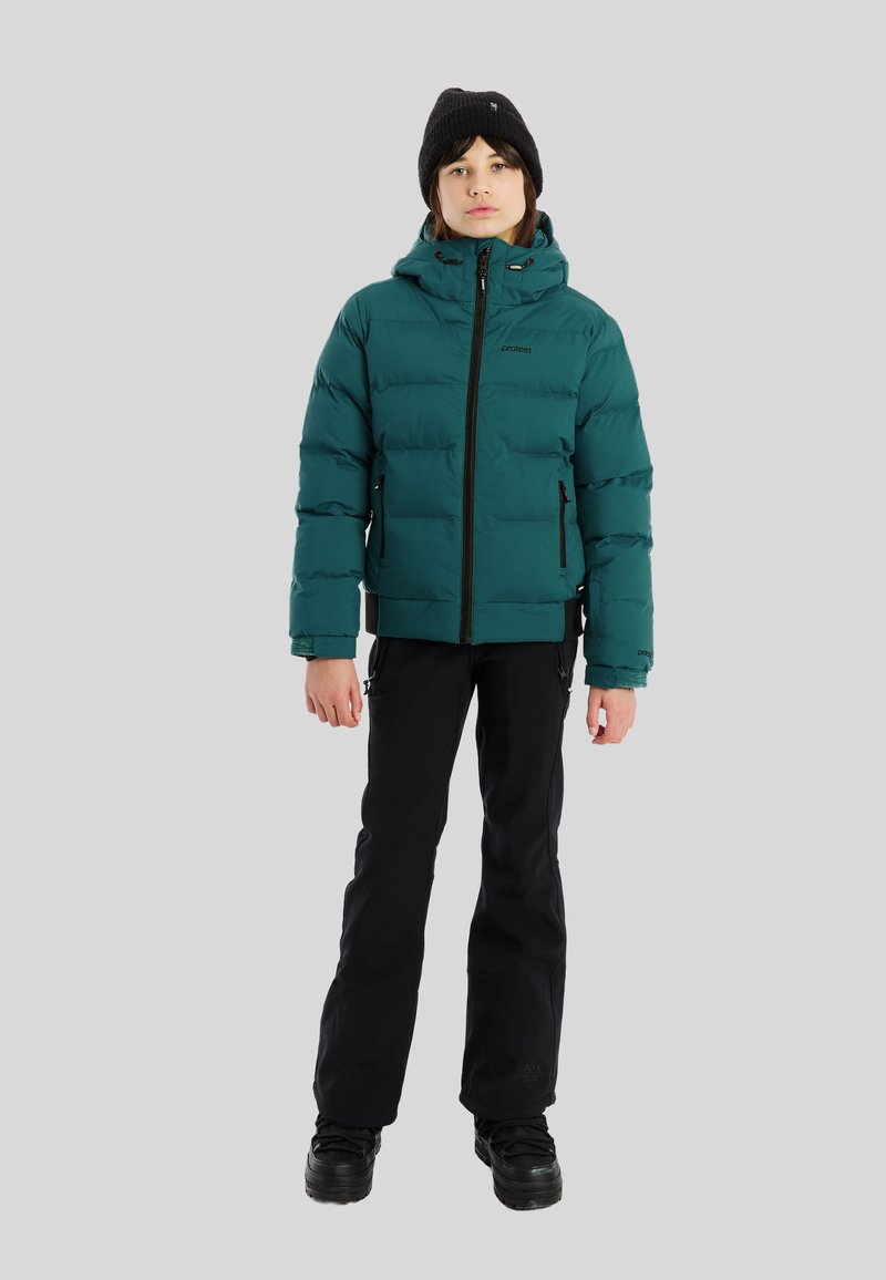 Jongere in een teal pufferjack, zwarte broek, zwarte beanie en zwarte laarzen, staande tegen een effen lichtgrijze achtergrond.