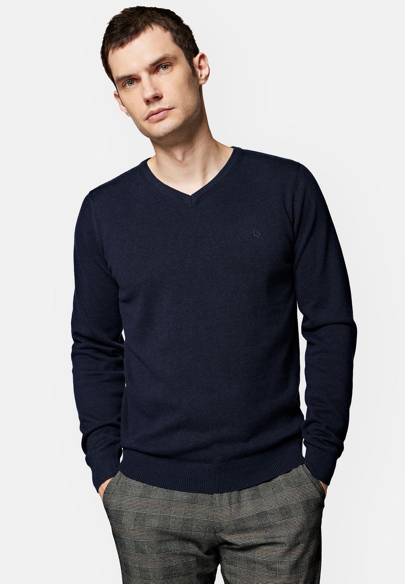 Granatowy sweter w serek z miękkiego materiału dzianinowego, z prążkowanymi mankietami i dołem, zestawiony z wzorzystymi szarymi spodniami w kratę.