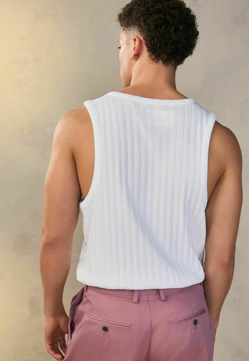 Weißes, geripptes Tanktop kombiniert mit hellrosanen Hosen, die über aufgesetzte Taschen und eine subtile Textur verfügen. Das Outfit zeigt eine lockere Passform.