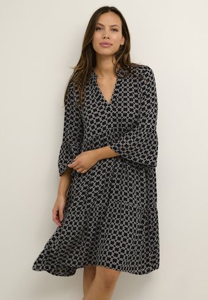 Eine Frau trägt ein schwarzes knielanges Kleid mit weißen geometrischen Mustern und Dreiviertelärmeln, steht vor einem schlichten hellen Hintergrund.