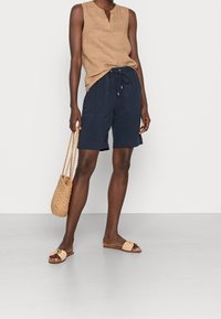 Haut sans manches beige, short à cordon de serrage bleu marine, sac panier tissé et sandales à enfiler marron clair, avec une texture de tissu lisse et une coupe décontractée.