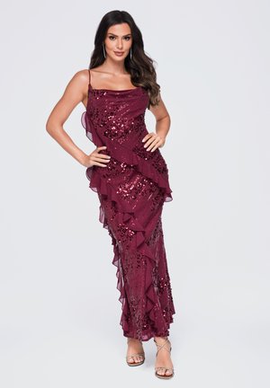Femme aux longs cheveux foncés portant une robe bordeaux sans manches à sequins avec volants, posant les mains sur les hanches avec des talons argentés à brides.