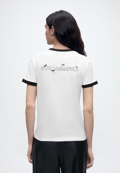 Жена с дълга тъмна коса носи бяла тениска с черна облицовка и текст "KARL LAGERFELD" и карикатурни кучета на гърба.