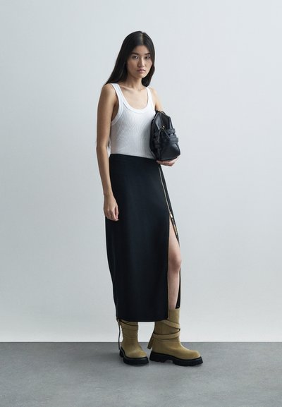 Kobieta w białym topie na ramiączkach, czarnej spódnicy z wysokim rozcięciem, beżowych botkach, trzymająca czarną torebkę na tle jasnym.