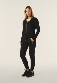 Sudadera negra con cremallera y un diseño ajustado, con un ligero brillo, combinada con leggins negros ajustados que presentan un patrón de huellas de patas en tono sobre tono en el costado.