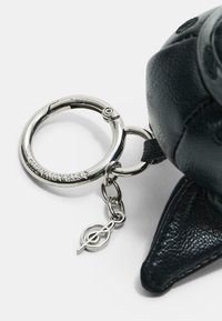 Dettaglio di una borsa in pelle nera con un portachiavi a forma di anello argentato, con il nome del marchio inciso, collegato da una short chain, e un piccolo charm con il logo.