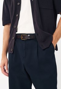 Black shirt with short sleeves, white t-shirt underneath, dark belt featuring a gold buckle, and dark pants with pleats.
