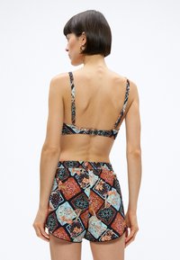 Γυναίκα με κοντά σκούρα μαλλιά που φορεί πολύχρωμο μπικίνι με μοτίβα και ταιριαστά σορτς, όρθια με την πλάτη στην κάμερα.