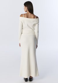 Maglione bianco off-the-shoulder con maniche lunghe abbinato a una gonna lunga in coordinato, caratterizzata da un motivo a maglia texturizzata. Scarpe nere con borchie.