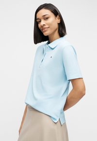 Γυναίκα με ίσια σκούρα μαλλιά που φοράει ανοιχτό μπλε πουκάμισο πόλο και μπεζ φούστα, στέκεται με τα χέρια πίσω από την πλάτη σε απλό φόντο.