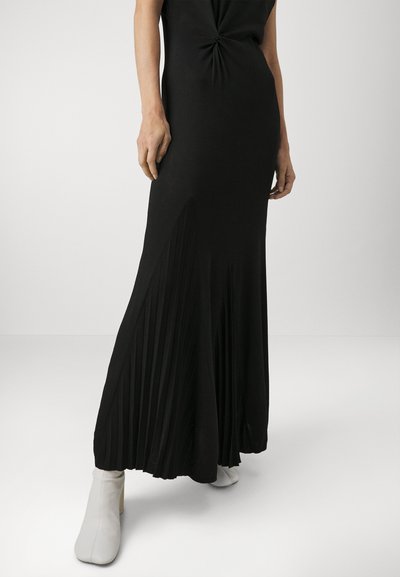 Rochie maxi neagră cu bust înnodat, având o fustă pliată. Țesătura pare netedă și elastică. Purtați-o cu cizme albe până la gleznă.