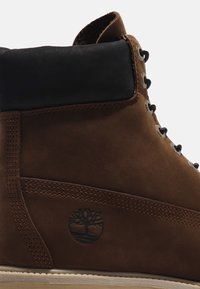 Botte en cuir marron avec col rembourré noir, œillets métalliques pour les lacets, couture visible et logo d'arbre embossé sur le côté.