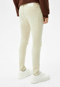 Jeans beige ajustados con un diseño desgastado, que cuentan con múltiples bolsillos y una etiqueta minimalista, combinados con zapatillas blancas.
