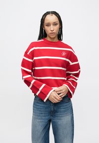 Haut rouge à manches longues avec des rayures horizontales blanches, col rond et manches larges. Présente un logo brodé sur le côté gauche de la poitrine. Associé à un jean bleu.