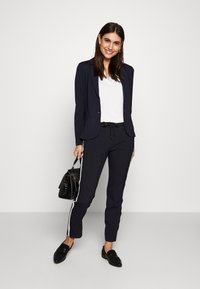 Blazer marine et joggers assortis avec des bandes latérales blanches. Haut blanc, mocassins noirs et un sac à main noir. Tissu lisse, coupe ajustée.