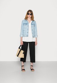 Jaqueta de denim azul clara sobre uma blusa branca, calças largas pretas, sandálias pretas e uma bolsa de palha trançada com detalhes pretos, óculos de sol usados.