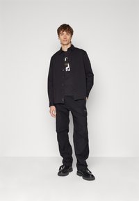 Camisa negra de botones sobre una camiseta gráfica negra, combinada con pantalones cargo negros y zapatos negros, todo en un diseño minimalista.