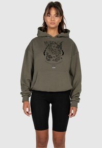Olive zielony, oversize'owy hoodie z kieszenią z przodu, wyposa�żony w czarny graficzny wzór kwiatowy oraz tekst „EST. 2015”. Noszony z czarnymi szortami motocyklowymi.