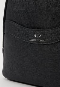 Mochila negra texturizada con un logotipo en relieve que dice "A|X ARMANI EXCHANGE" en color plateado. El diseño incluye un acento cosido y detalles de cremallera.