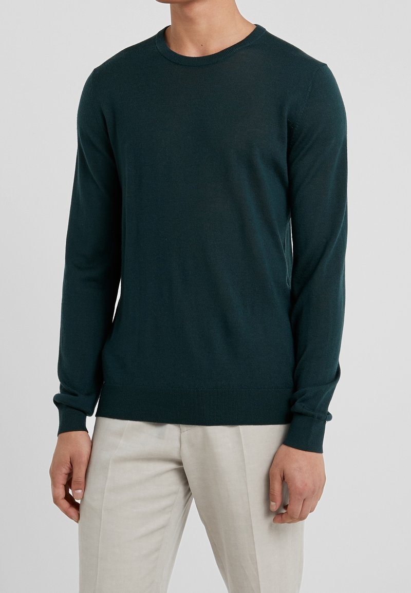 Ciemnozielony sweter z okrągłym dekoltem, wykonany z miękkiego materiału, z długimi rękawami i ściągaczami przy mankietach. Nosi się go na jasnych spodniach.