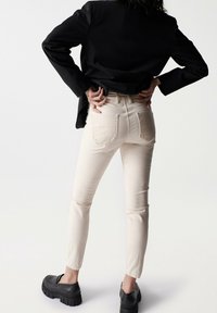 Jeans beige ajustados con cintura alta, que presentan bolsillos traseros texturizados. Combinados con una americana negra y unos mocasines negros gruesos.