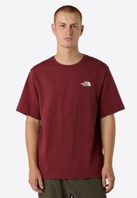Camiseta de algodón burdeos con mangas cortas, cuello redondo y un logo blanco en el lado izquierdo del pecho. Ajuste casual y textura suave.
