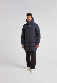 Giacca imbottita di colore blu navy con cappuccio, stretto in vita, dotata di zip completa, ampie sezioni trapuntate e un logo sul petto.