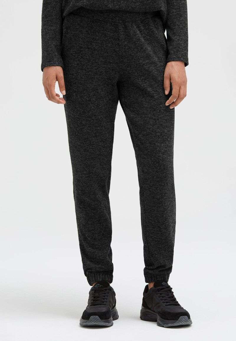 Dunkelgraue Jogginghosen aus weichem Stoff mit elastischem Bund und Bündchen an den Knöcheln, kombiniert mit schwarzen Sportschuhen.