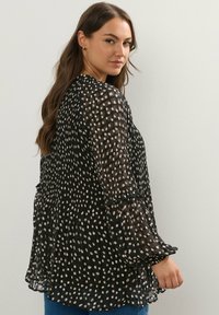 Svart långärmad topp med en flödande design, med en plisserad textur och vita prickar. Fitted manschetter med volangdetaljer.