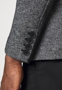 Grijze blazerstof met een gestructureerd patroon, met vier zwarte knopen op de manchetten. Arm en zwarte broek gedeeltelijk zichtbaar.