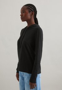 Zwarte hoodie met lange mouwen en een relaxte pasvorm, met een trekkoord bij de hals, zachte textuur, gecombineerd met lichtblauwe jeans, bekeken vanaf de zijkant.