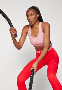 Roze sportski grudnjak i crvene tajice s bijelim Nike logotipima. Model drži debelu, crnu borbenu uže, prikazujući dinamičnu vježbu.