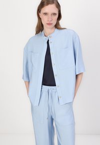 Camisa de botón azul claro con mangas cortas, dos bolsillos en el pecho y un cuello redondeado. Llevada sobre una blusa oscura, combinada con pantalones a juego.