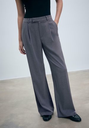 Persona che indossa pantaloni grigi a vita alta e a gamba larga con pieghe, top nero, una mano in tasca, in piedi su un pavimento in cemento.