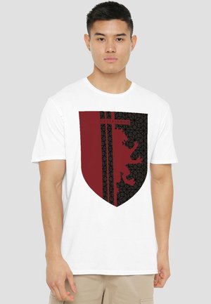 Young man wearing white t-shirt with maroon and black Gryffindor shield graphic on front against plain background.