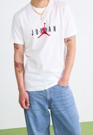 Person, der ein weißes Jordan-T-Shirt mit rotem Logo, helle blaue Jeans, eine goldene Kettenhalskette mit dem Buchstaben "A" und bunte Armtätowierungen trägt.