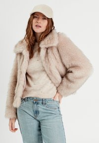 Veste en fausse fourrure de couleur rose pâle avec un col cranté, associée à un pull beige avec des accents de perles et un jean bleu clair.