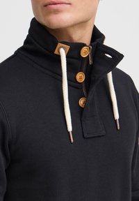 Sudadera negra con cuello alto, que cuenta con cordones en color crema, botones de madera y una parte delantera con cremallera. Tela texturizada visible.
