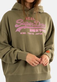 Olijfgroene oversized hoodie met een glinsterend roze "SuperDry"-logo, ribgebreide manchetten en een oranje labeldetail op de mouw. Zachte, gestructureerde stof.